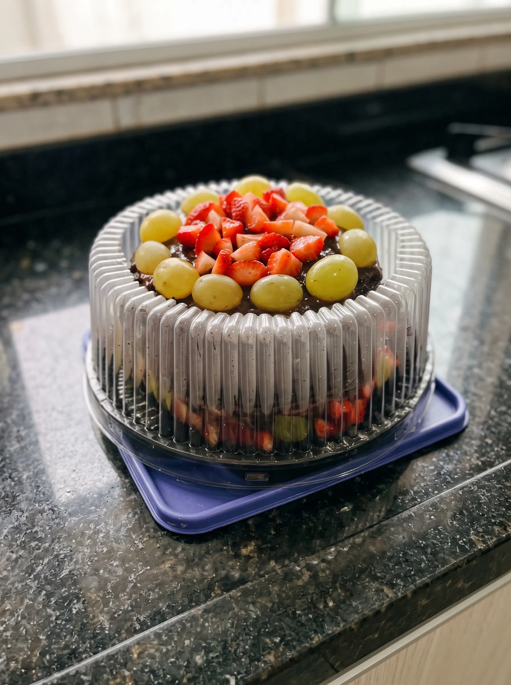 Bolo de chocolate coberto de morangos e uvas, numa embalagem transparente